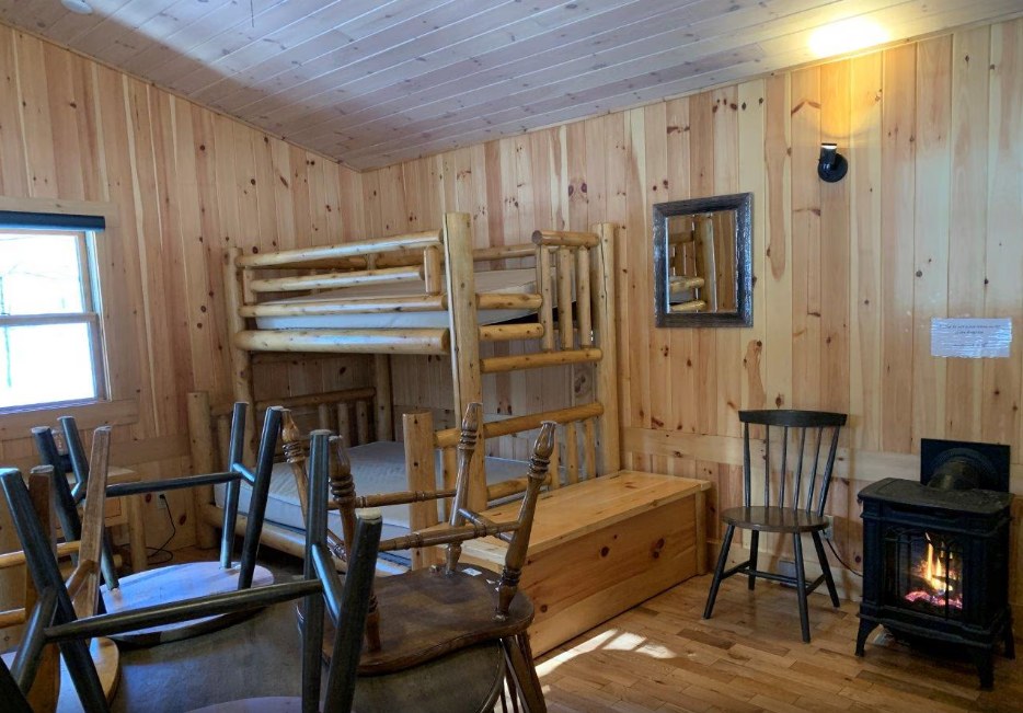 Inside an Arrowhead rustic cabin: knotty-pine walls and ceiling, a log bunk bed with a double bottom bunk and single top, a black propane fireplace burning in the corner, and a table with chairs stacked on it.