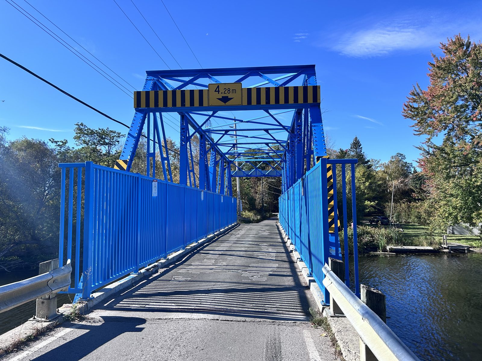Looking down the deck of a one-lane steel truss bridge painted bright blue, a yellow 4.28 m clearance sign across the top girder, river on the right