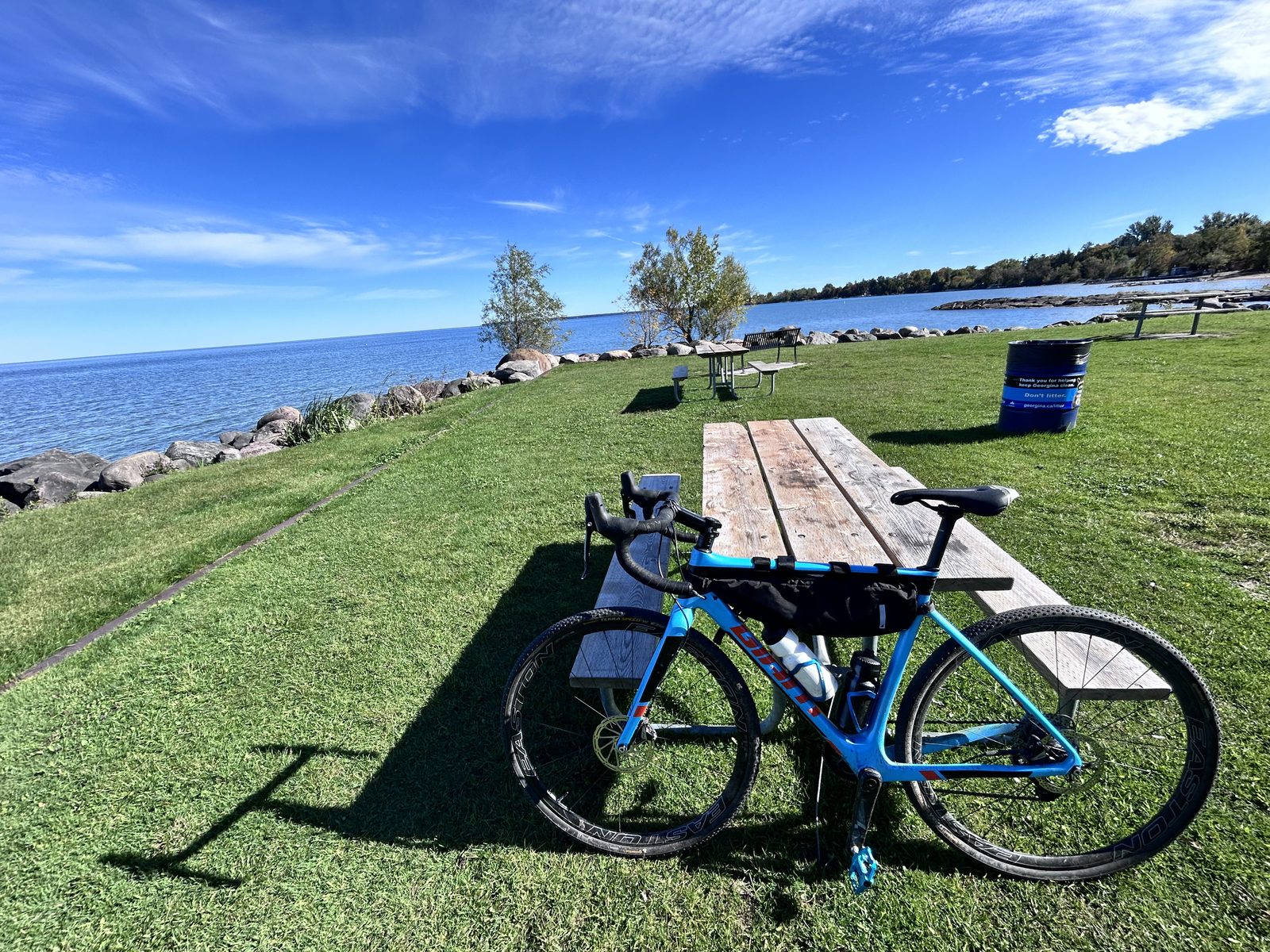 A blue gravel bike leaning on a picnic table on mown grass at the edge of Lake Simcoe, boulder shoreline and open blue water behind