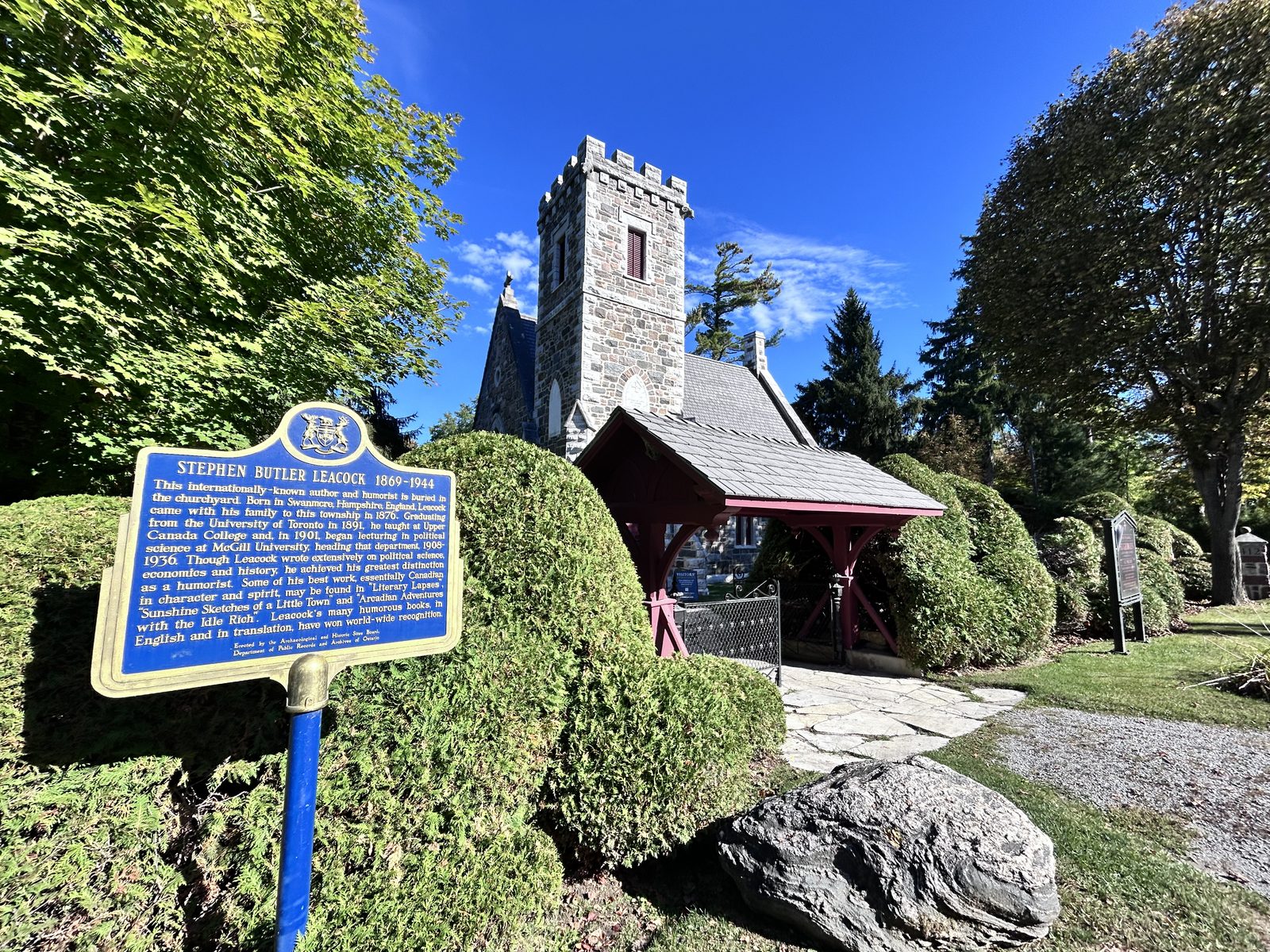 A small grey stone church with a square crenellated tower behind clipped cedar hedges, a blue Ontario heritage plaque for Stephen Butler Leacock in the foreground, a timber lychgate to the right